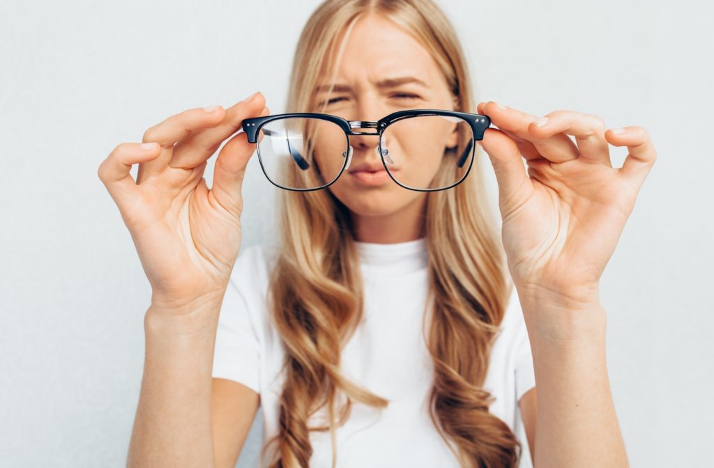 A woman holding out a pair of glasses while she squints attempting to look through the lenses