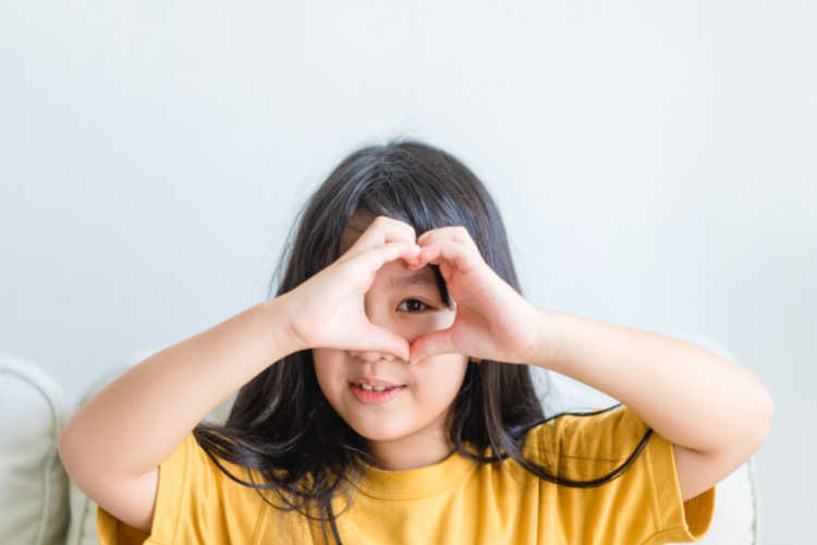 A young girl making a heart shape with her hands around her left eye