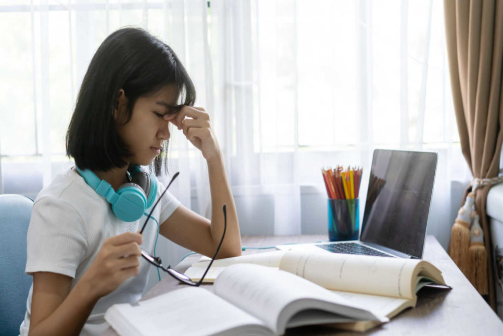 A young girl doing homework holding her glasses in one hand, rubbing her eyes with the other due to problems with her eyesight