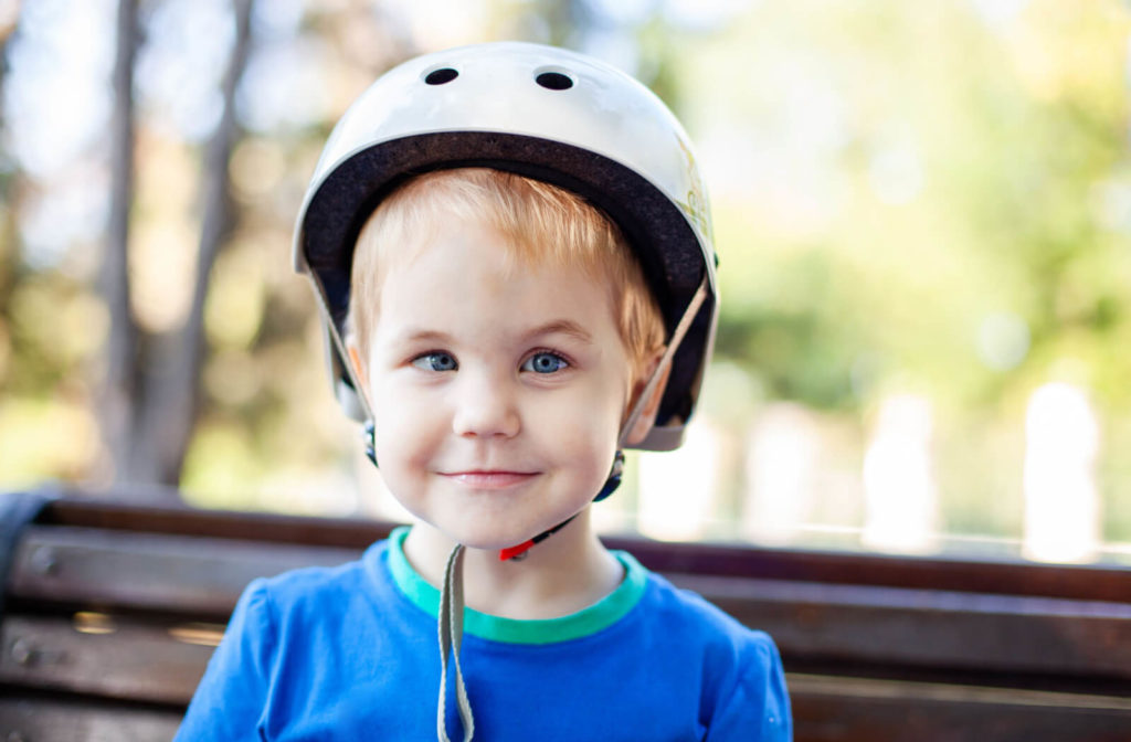 A boy in a blue shirt and wearing a white coloured helmet outside has a crossed eye,  a condition in which one eye does not point in the same direction as the other eye.