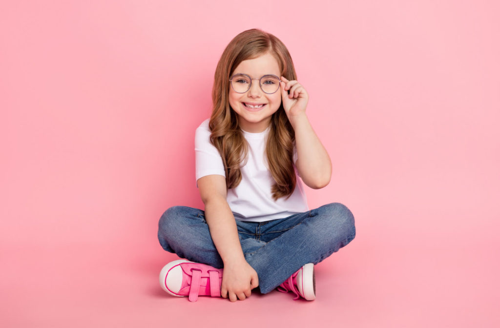 Young girl against a pink background holding her glasses up with her hand.