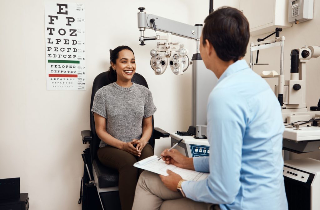 A woman sitting in an eye exam room, smiling at her eye doctor with an eye chart behind her and a phoropter beside her.