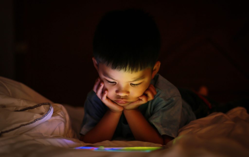 A young child lays on their stomach and watches a brightly colored tablet on the floor in front of them in the dark.