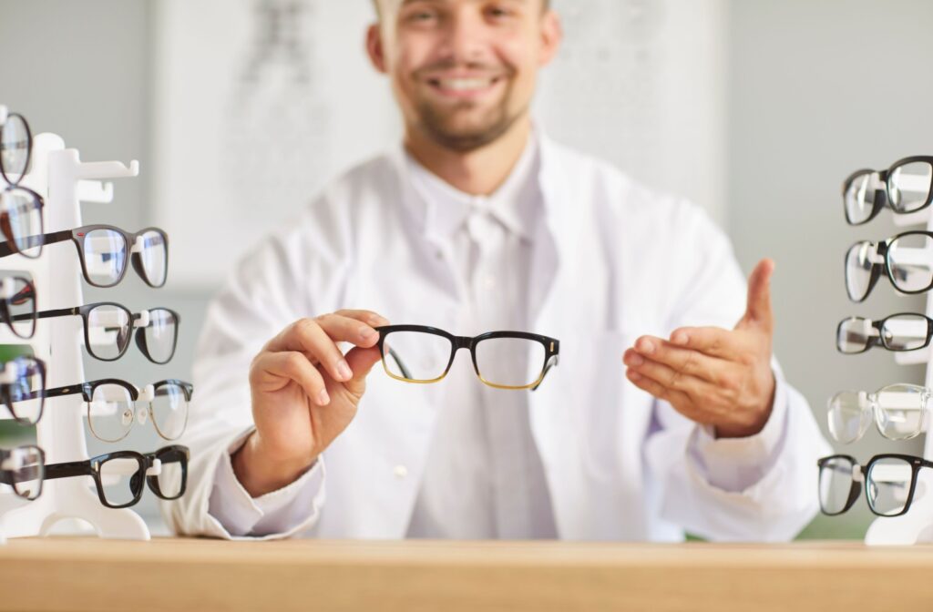 Optician in white coat holding eyeglasses with display racks of frames on either side