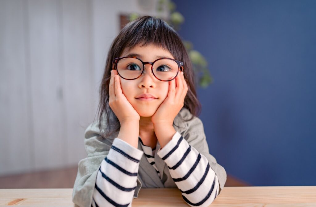 Young girl with glasses rests her face in her hands, looking thoughtful, illustrating how amblyopia may affect a child’s confidence and visual development.