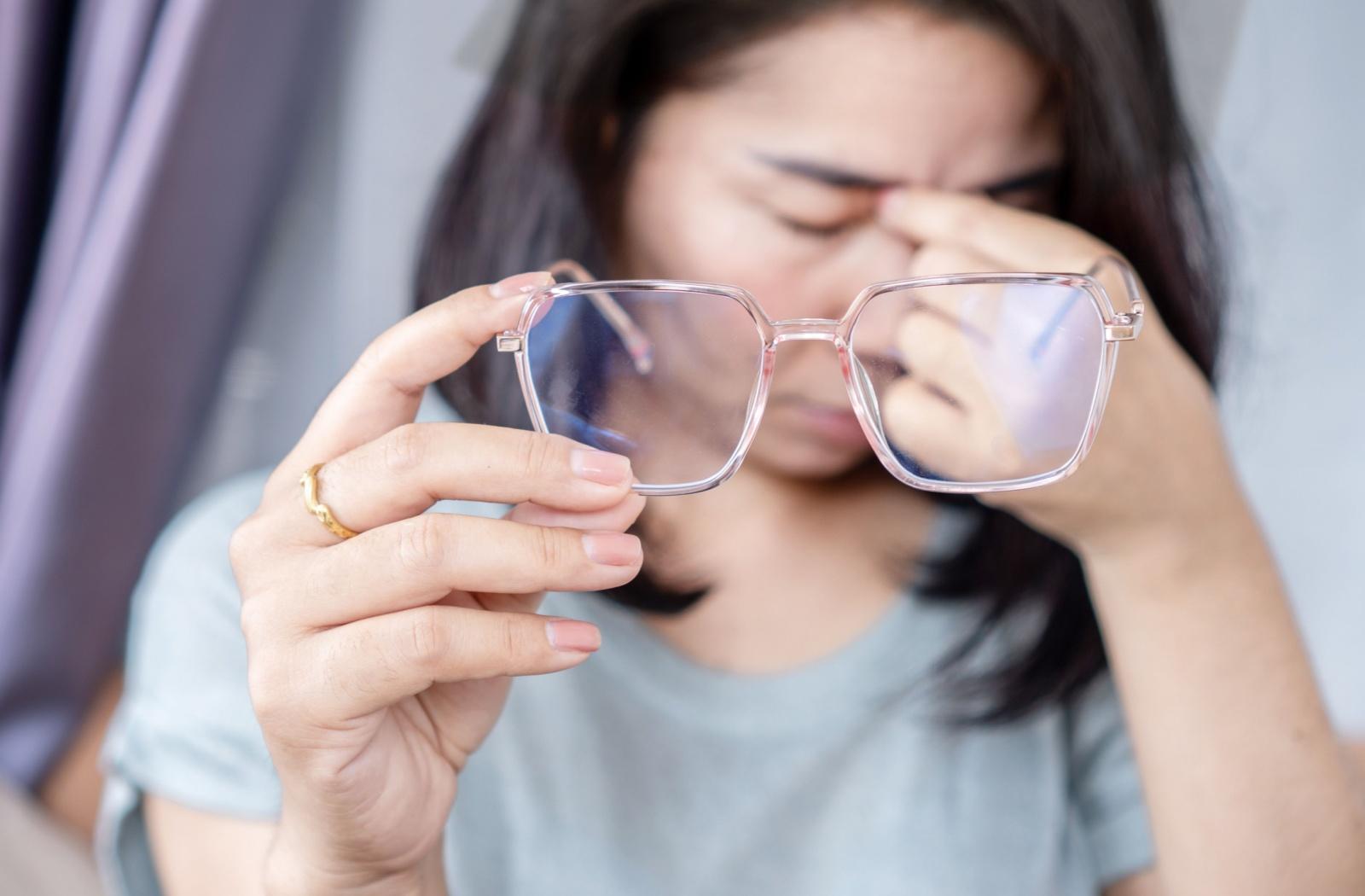 A person rubbing in between their eyes and holding their glasses up to the camera